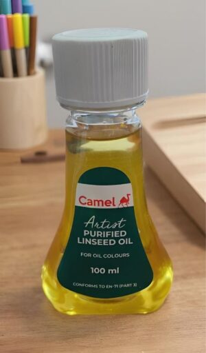 Camel Artist Purified Linseed Oil bottle, 100 ml, on a wooden table with colored pencils in the blurred background.