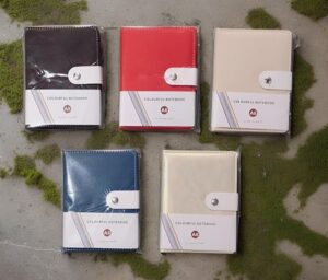 Five A6 colorful notebooks in plastic wrap with white straps, arranged on a mossy gray surface (black, red, beige, blue, cream).