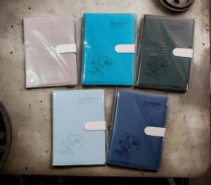 Five pastel-colored hardcover notebooks/planners with white straps laid on a concrete surface (pink, turquoise, gray, light blue, navy).