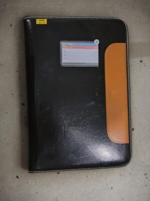 Black textured binder with an orange vertical pocket on the right and a small transparent ID card slot near the top left, placed on a concrete floor