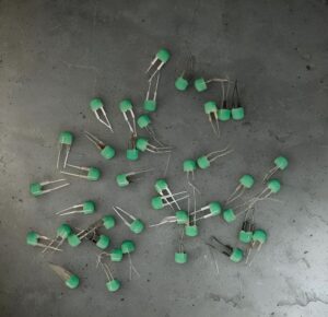 Scattered green epoxy-coated radial capacitors with two leads on a dark gray surface.