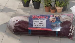 Rolled bundle of maroon shuttle net cotton Rajan packed in plastic, placed on a tiled patio with potted plants in the background.