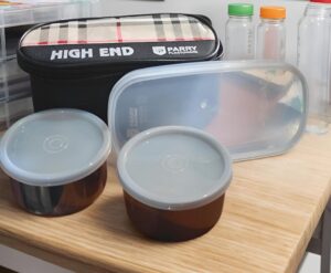 Two brown glass-like meal containers with gray lids sit on a wooden table, with a black High End lunch bag in the background.