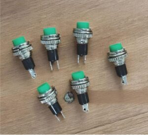 Six green push-button switches with metal collars on a wooden surface laid out for display.