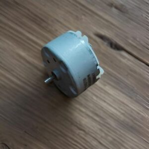 Small gray plug‑in electrical adapter lying on a wooden floor with a metal pin protruding from the side.
