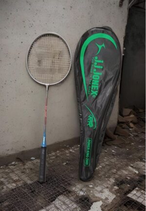 Badminton racket leaning against a concrete wall beside a black-green Power King 2600 racket bag.