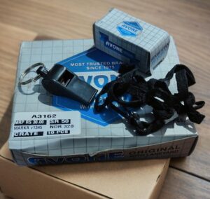 Close-up of Avon-branded packaging with a keychain tool and tangled black strap resting on a gray grid-patterned box.