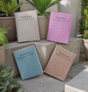 Four glittery mood diaries in pastel colors (blue, beige, pink, gray) arranged on stone steps with potted plants, each cover reads 'Mood diary'.