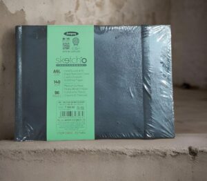Black hardbound sketchbook in plastic wrap on a shelf with a mint-green label reading 'sketcho' and product specs (A6L, 96 pages).