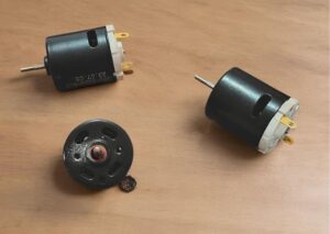 Two small brushed DC motors with yellow terminal tabs lie on a wooden surface beside a black circular end cap component with a red center bearing and multiple ventilation slots.