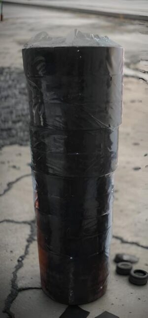 Tall black cylinder wrapped in duct tape standing on a cracked concrete surface; small rubber rings lie nearby on the ground.