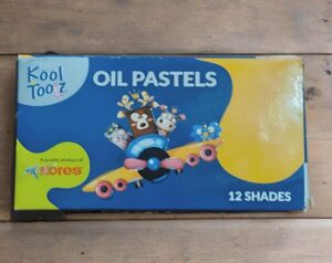 Blue and yellow oil pastels box labeled 'Oil Pastels' with '12 Shades', featuring a cartoon airplane carrying animal characters in the center (Kores brand).