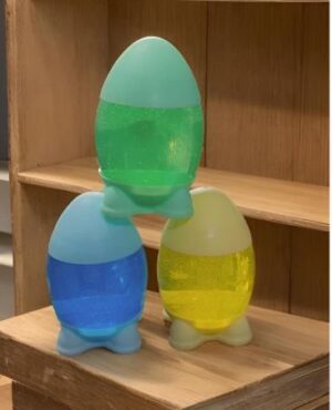 Three pastel-colored, egg-shaped water toys with blue feet, stacked on a wooden shelf: a large green egg on top, a blue egg on the left, and a yellow egg on the right.