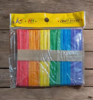 Colorful rainbow craft sticks in a plastic bag with a yellow header labeled 'JC DIY Craft Sticks' on a wooden surface.