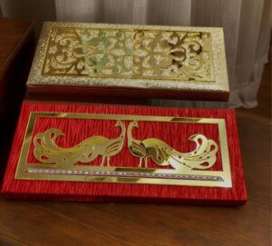 Two decorative boxes with gold designs, stacked on a wooden surface: a gold filigree lid over a textured red box and a matching gold panel on top.
