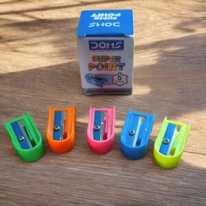 Set of five colorful pencil sharpeners (green, orange, pink, blue, yellow) on a wooden surface with a Super Point 5 pieces box in the background.