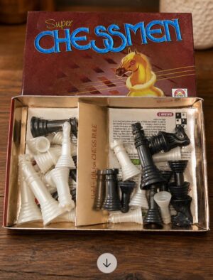 Open box of a vintage 'Super Chessmen' game with white and black plastic chess pieces spilling from a gold tray inside a brown-red box showing a horse image.