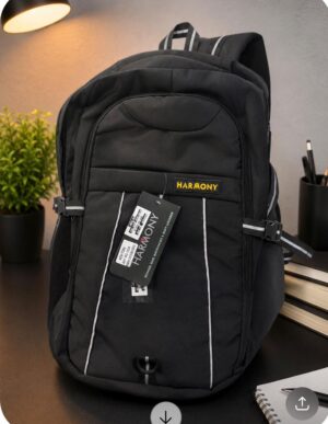 Black Harmony backpack with white piping and a tag, placed on a desk beside a notebook and plant.