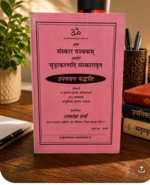 Pink book with Devanagari text on a wooden desk, framed by a pen and notebook on left and stacked books on the right.
