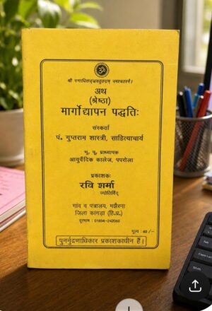 Yellow book cover with black Devanagari text sits on a wooden desk, next to a pen holder and a laptop keyboard in the background.