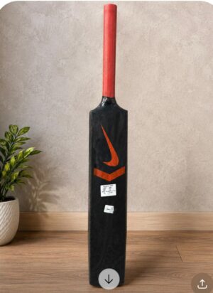 Tall black cricket bat with a red grip standing upright on a wooden floor near a potted plant.