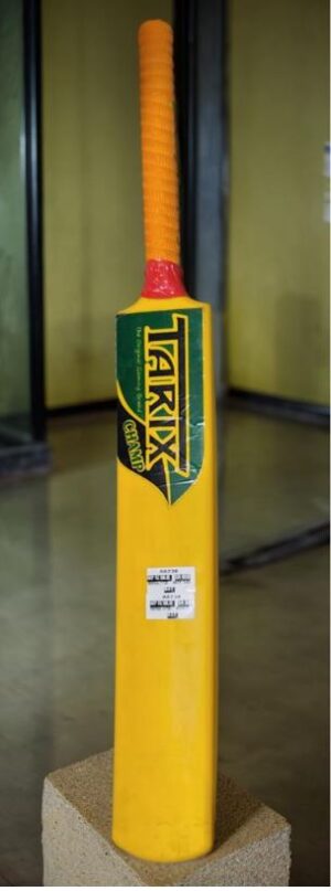 Bright yellow cricket bat with an orange grip mounted on a display pedestal.