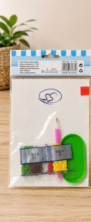 Clear view of a bead craft kit still in its plastic packaging on a wooden table, showing color beads, a green tray, and a pink tool pen.