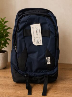 Navy blue backpack with black panels on a wooden surface, tag attached to the top handle and a plant in the background.