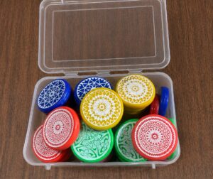 Clear plastic box holding round, colorful lids with white mandala patterns (blue, red, yellow, green) on a wooden surface outside the container.