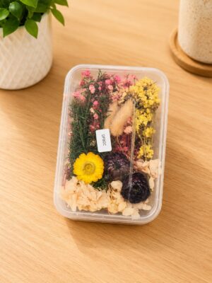 Clear plastic container holding an arrangement of dried flowers in yellow, pink, and purple on a wooden surface.