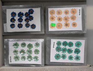 Four display sheets each with daisy-like flower stickers in blue, orange, and green tones arranged in a 2x2 grid, on a gray background.”,