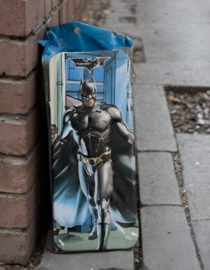 Tall Batman-themed rectangular box leaning against a brick wall on a sidewalk, with a blue plastic cover on top and a cityscape background in the artwork.