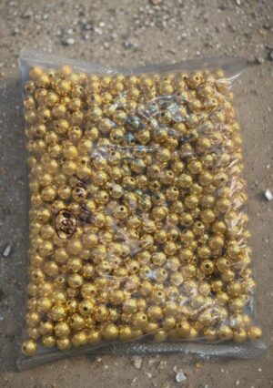 Transparent plastic bag filled with many small shiny gold beads, resting on a rough concrete surface.