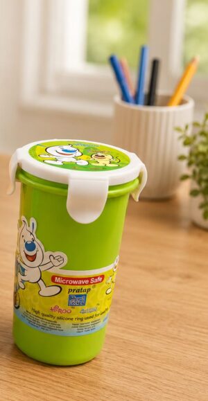 Green kids' lunch tumbler with white lid and cartoon characters sits on a wooden desk, label reads 'Microwave Safe'. In the blurred background, a cup of colored pens sits near a window.