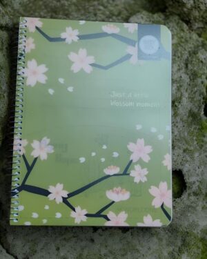 Green spiral notebook with pink cherry blossoms on a mossy surface; cover reads 'Just a little blossom moment'.