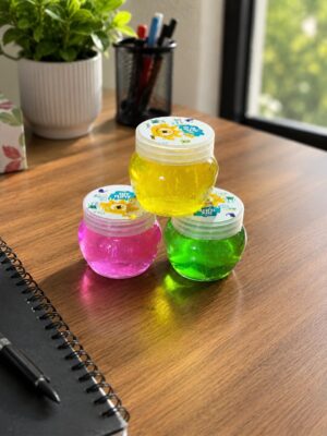 Three small, translucent slime jars in yellow, pink, and green arranged on a wooden desk with a notebook nearby and a plant in the background (indoor scene).