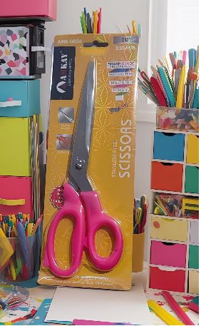 Pink-handled scissors resting against yellow packaging on a colorful craft desk with bright organizers and paper supplies nearby.
