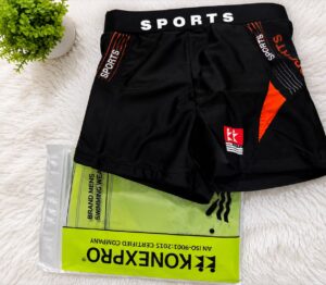 Black athletic shorts with orange panels and a white 'SPORTS' waistband laid over a neon-green packaging bag on a white rug.