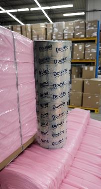 Industrial warehouse scene with pink insulation sheets on pallets and a tall white roll standing upright in the foreground, with cardboard boxes on shelves in the background.
