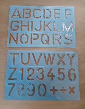 Blue stencil boards with copper cutouts showing the full uppercase alphabet and digits 0–9 on a wooden surface.