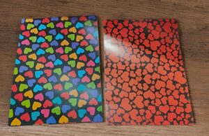Two notebooks on a wooden table; left one has multicolored hearts on black, right one has red hearts on a dark background.