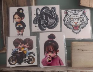 Five cartoon art prints displayed on a shelf: girl with rainbow sweater, black abstract creature, snarling tiger, two girls on a motorcycle, and a girl pointing forward.