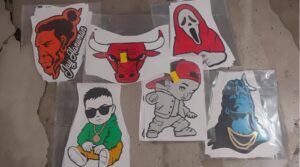 Six cartoon-style vinyl stickers laid on a concrete floor: an angry red-haired man, a red bull, a ghost in a red hood, a cool kid in sunglasses, a kid in a cap, and a blue statue bust with gold chains.
