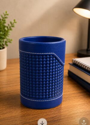 Blue cylindrical leather desk organizer with white stitching on a wooden desk, flanked by a plant and a lamp