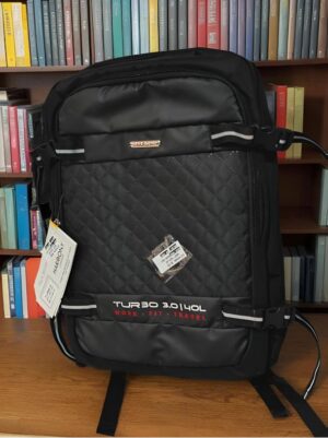 Black quilted backpack with multiple straps and tags, placed on a wooden surface in front of colorful bookshelves.