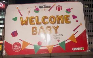 Bright welcome sign reading 'WELCOME BABY' in gold-letter balloons, decorated with cupcakes, gifts, candles, and a colorful pennant banner; set against a white background with a red base.