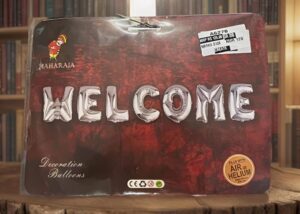Package for 'WELCOME' balloon letters on a dark red background with 'Decoration Balloons' text and helium/Air stamp, on a wooden surface.