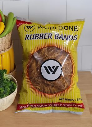 Yellow World One Rubber Bands package on a wooden counter; clear window showing rubber bands, brand logo in center, contains 50g net weight.