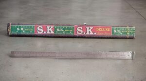 Metal ruler lying on a concrete floor with its case above showing 'S.K. Deluxe Steel Scale' text.