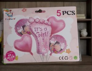 Box of five pink baby-shower balloons featuring a central foot-shaped balloon with 'It's a Girl!' text, surrounded by heart-shaped balloons on a pink packaging design.